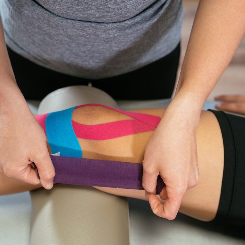 Photo detail of the hands of a physiotherapist woman gluing purple medical tape on another celestial tape and another pink one on the knee of a patient. Concept of muscle health and relaxation. Close-up